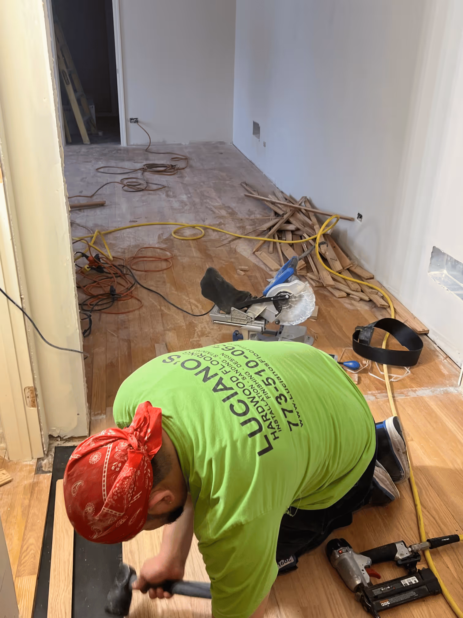 Worker with red bandana kneeling to install hardwood flooring using a hammer in a room under renovation.