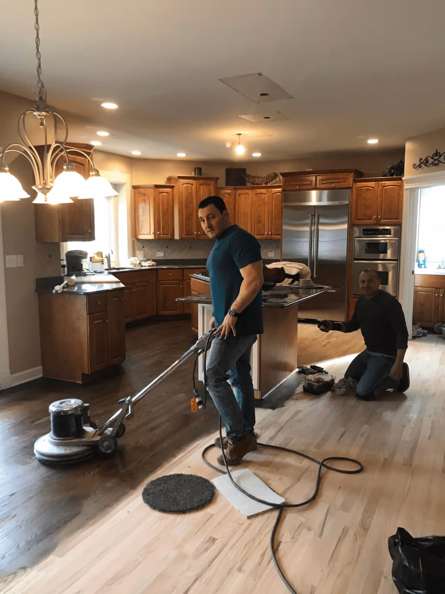Two men refinishing a hardwood floor in a kitchen, one using a floor sander, the other kneeling.