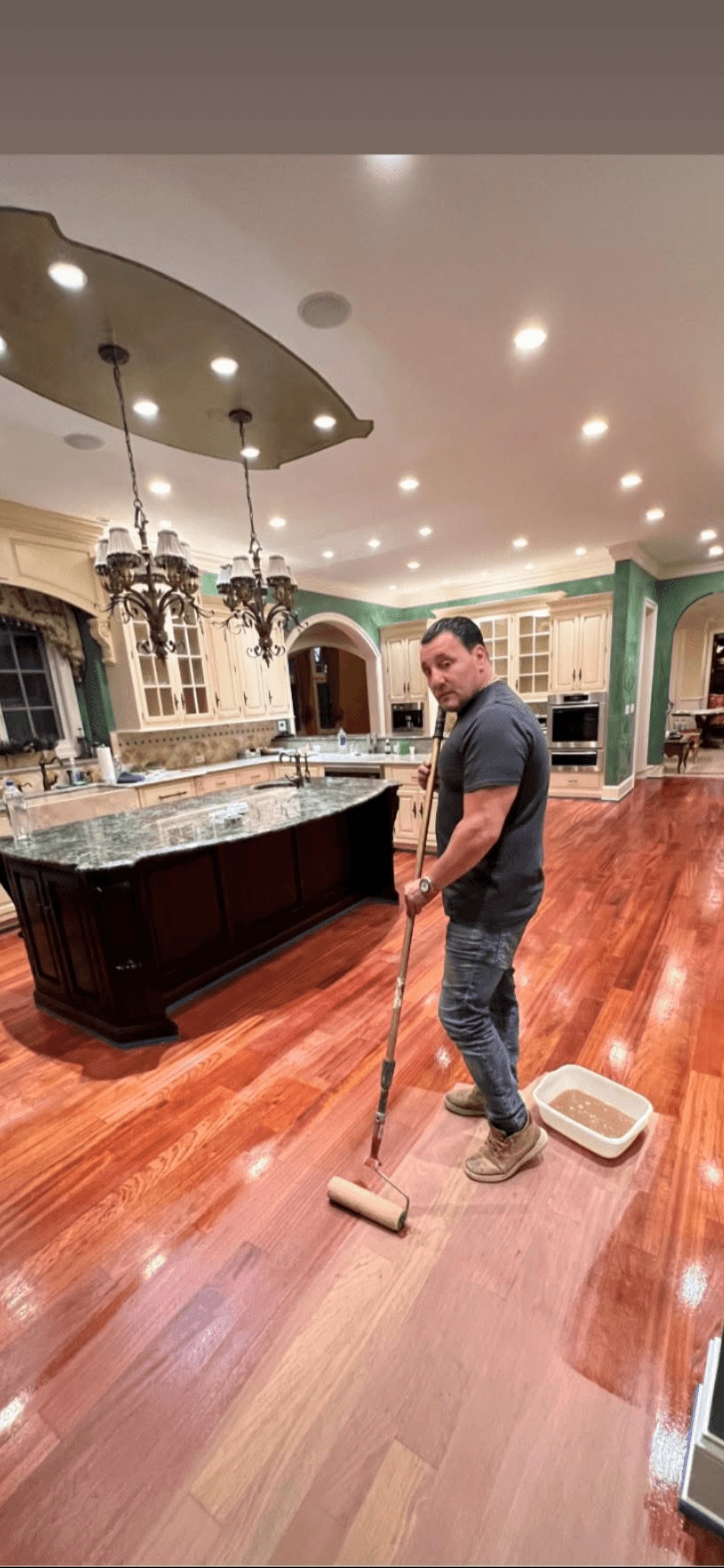 Person applying finish to hardwood floor in spacious, two-story living room with staircase and fireplace.