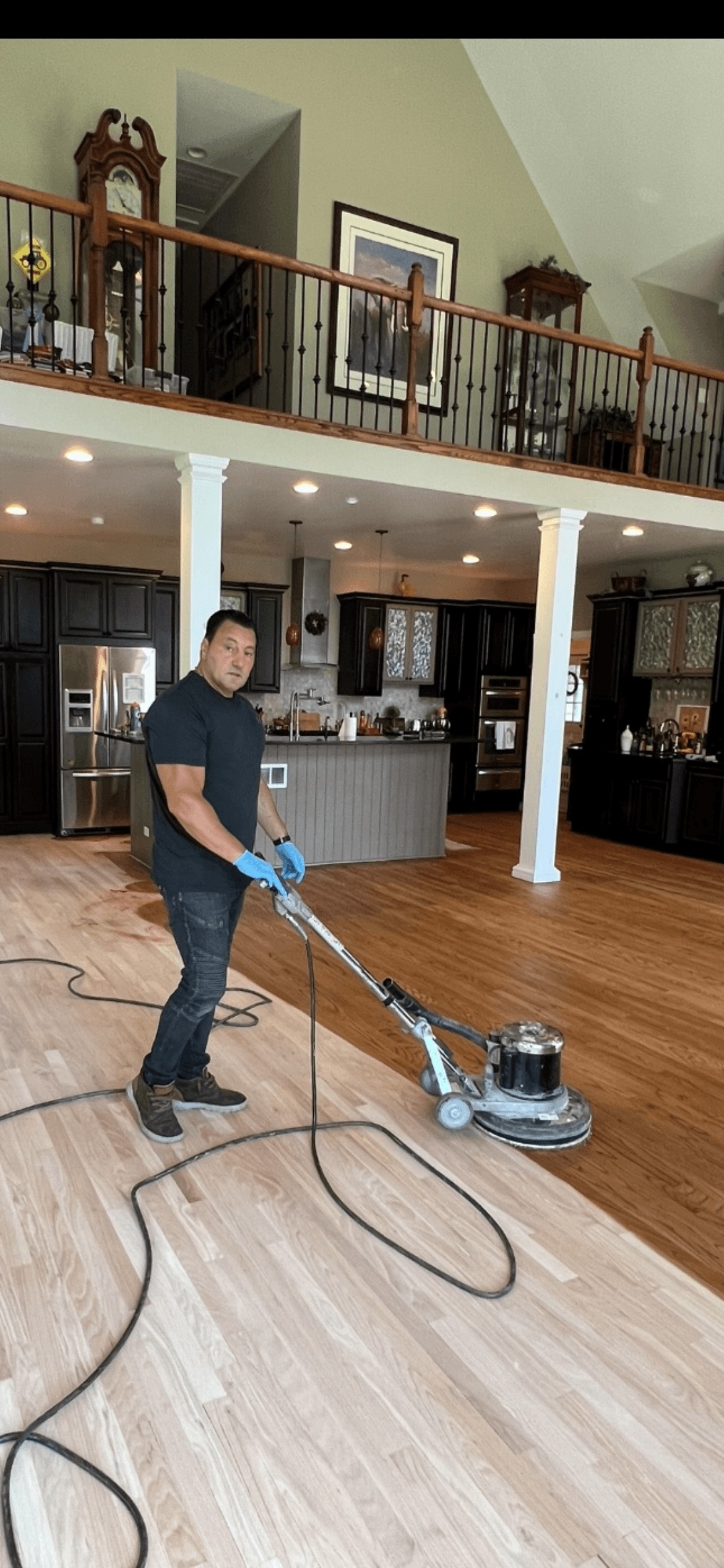Man using floor buffer to polish wooden floor in a large kitchen with dark cabinets.