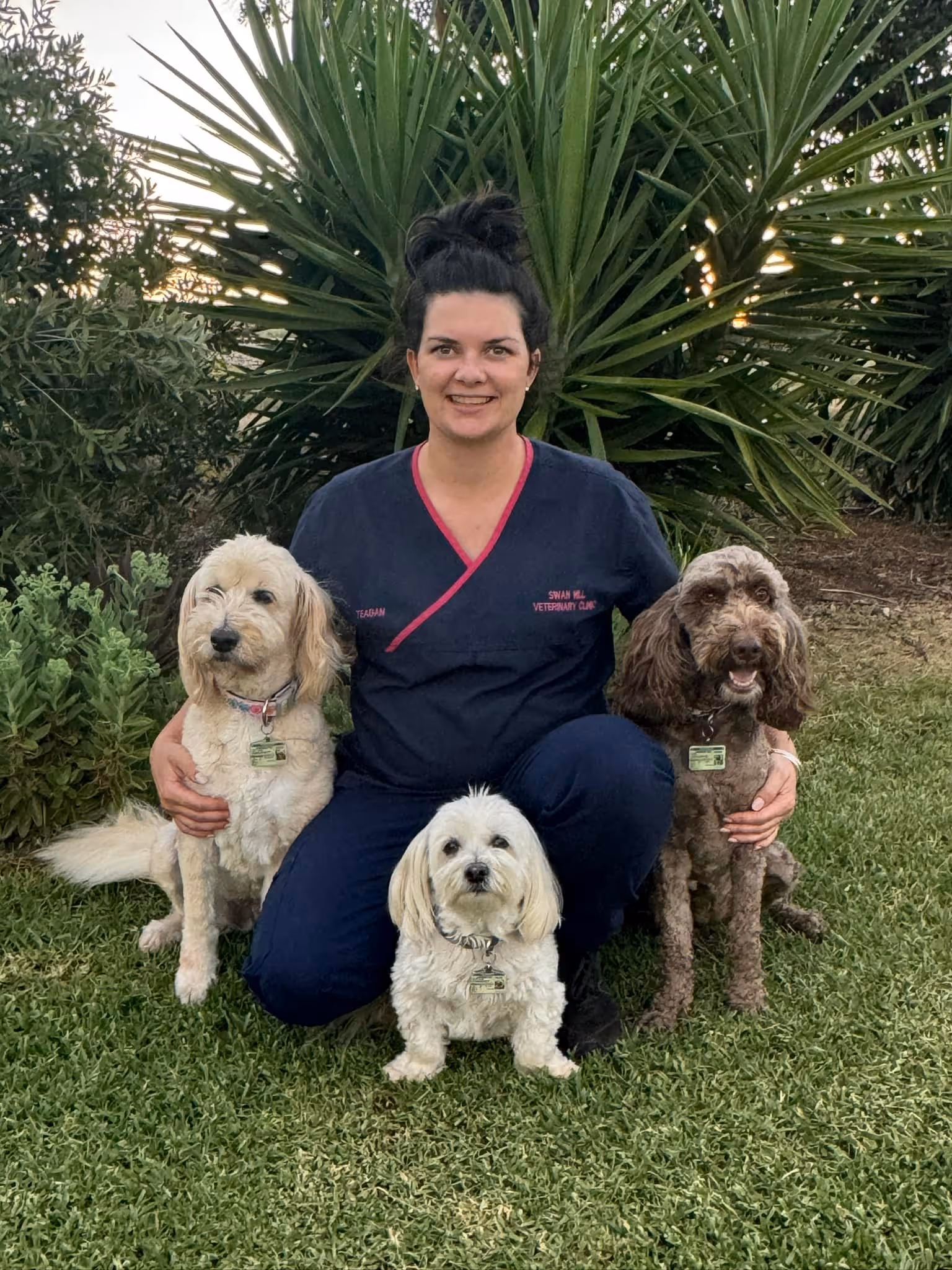 Woman in navy scrubs kneeling on grass outdoors with three small dogs, two sitting beside her and one in front.