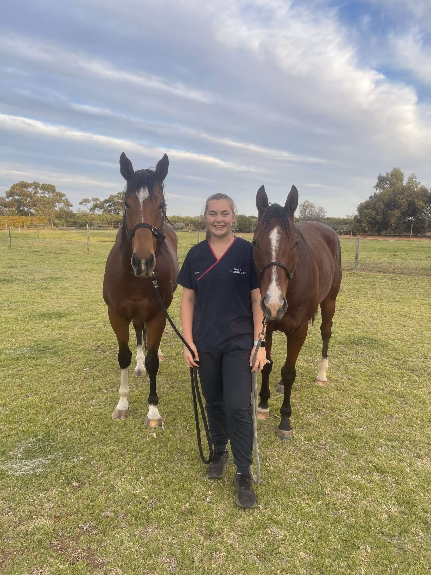 Smiling woman in navy scrubs walking two brown horses on a grassy field under a partly cloudy sky.