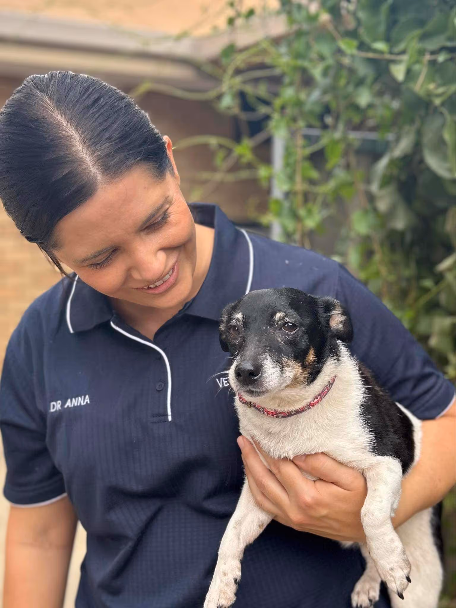 Smiling woman in a navy polo shirt holding a small black and white dog outdoors.
