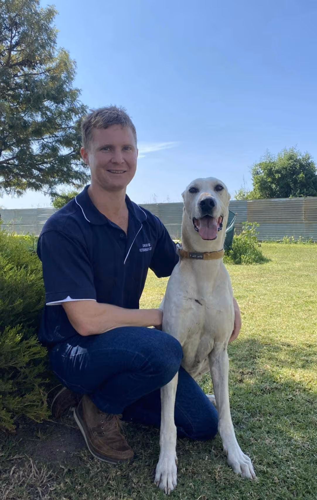 Man in navy scrubs kneeling on grass and smiling next to a large white dog sitting beside him.