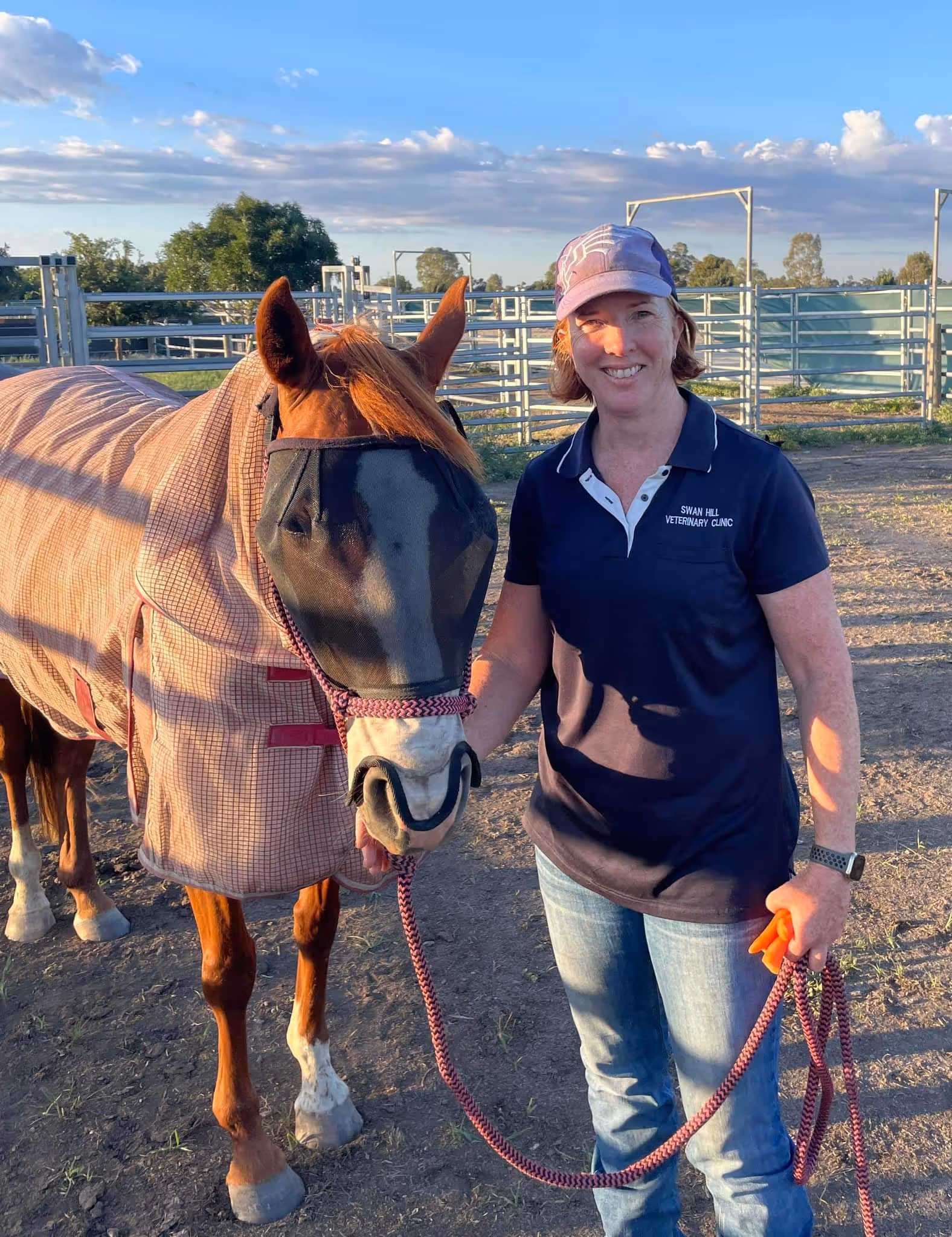Woman in a baseball cap and polo shirt holding the reins of a brown horse wearing a protective fly mask and blanket.