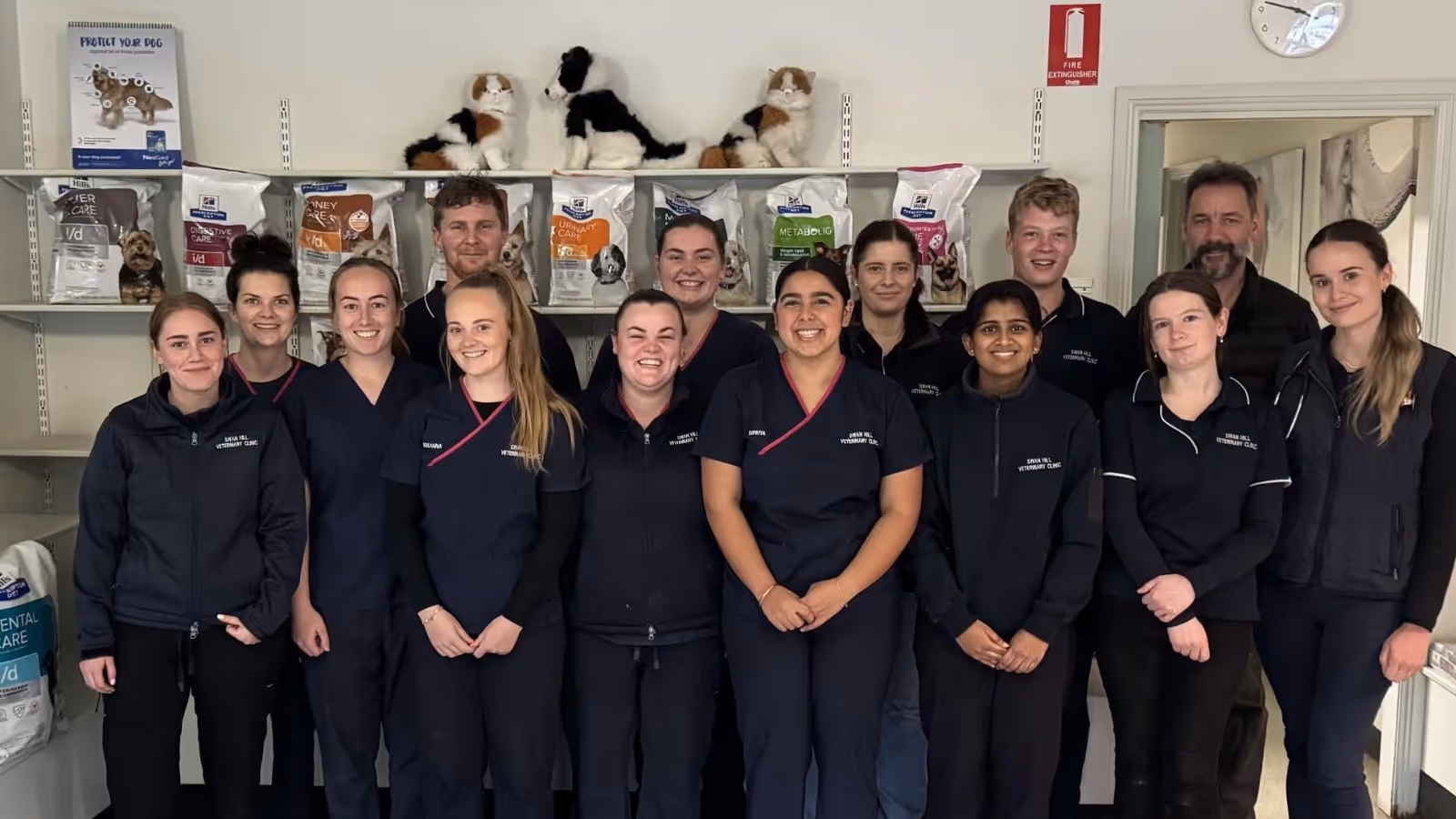Group photo of thirteen people standing indoors, all dressed in dark casual attire, with shelves and stuffed dog toys on the wall behind them.