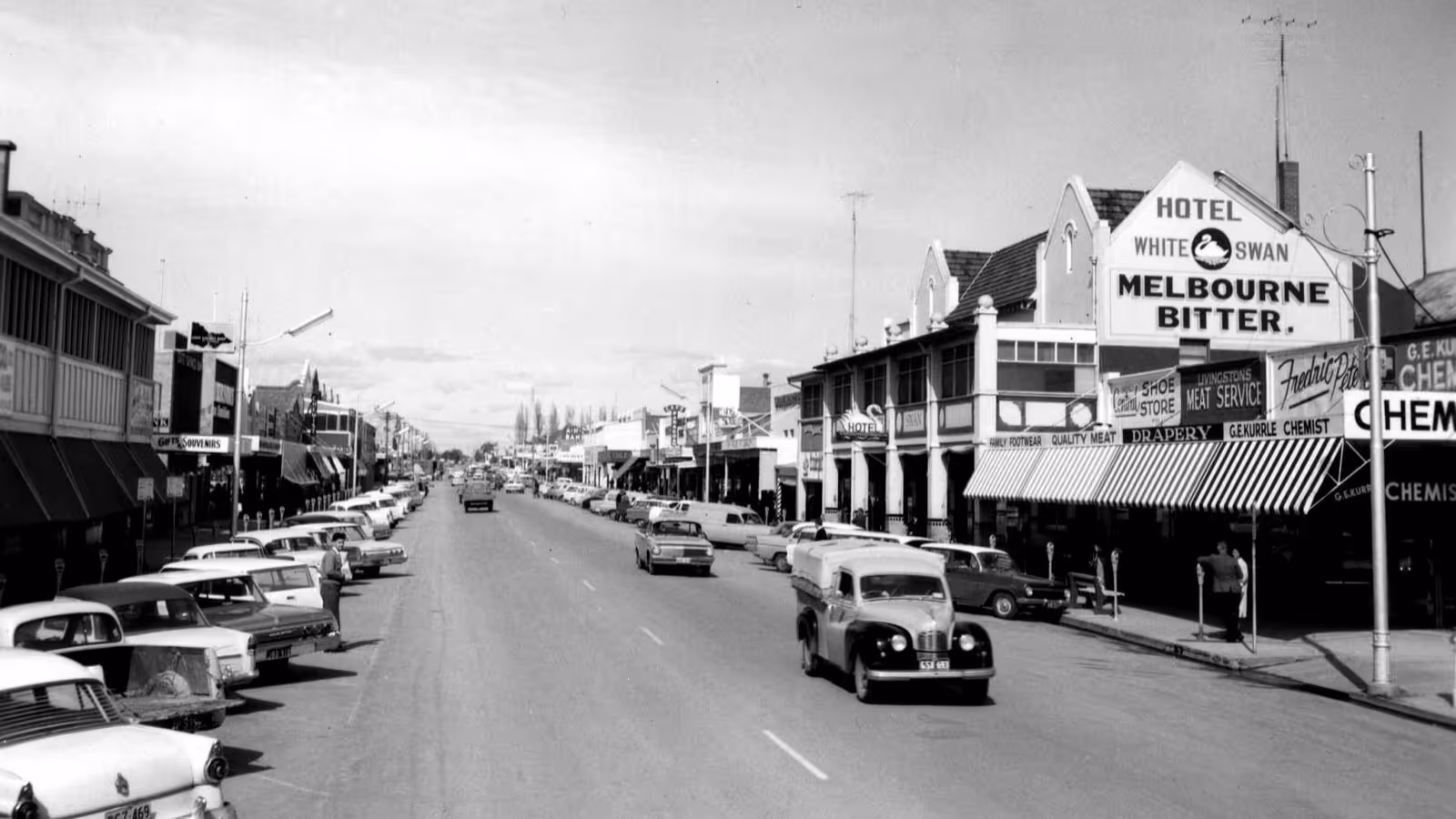 Black and white street view of a town with vintage cars parked along both sides and a hotel sign reading 'White Swan Melbourne Bitter'.