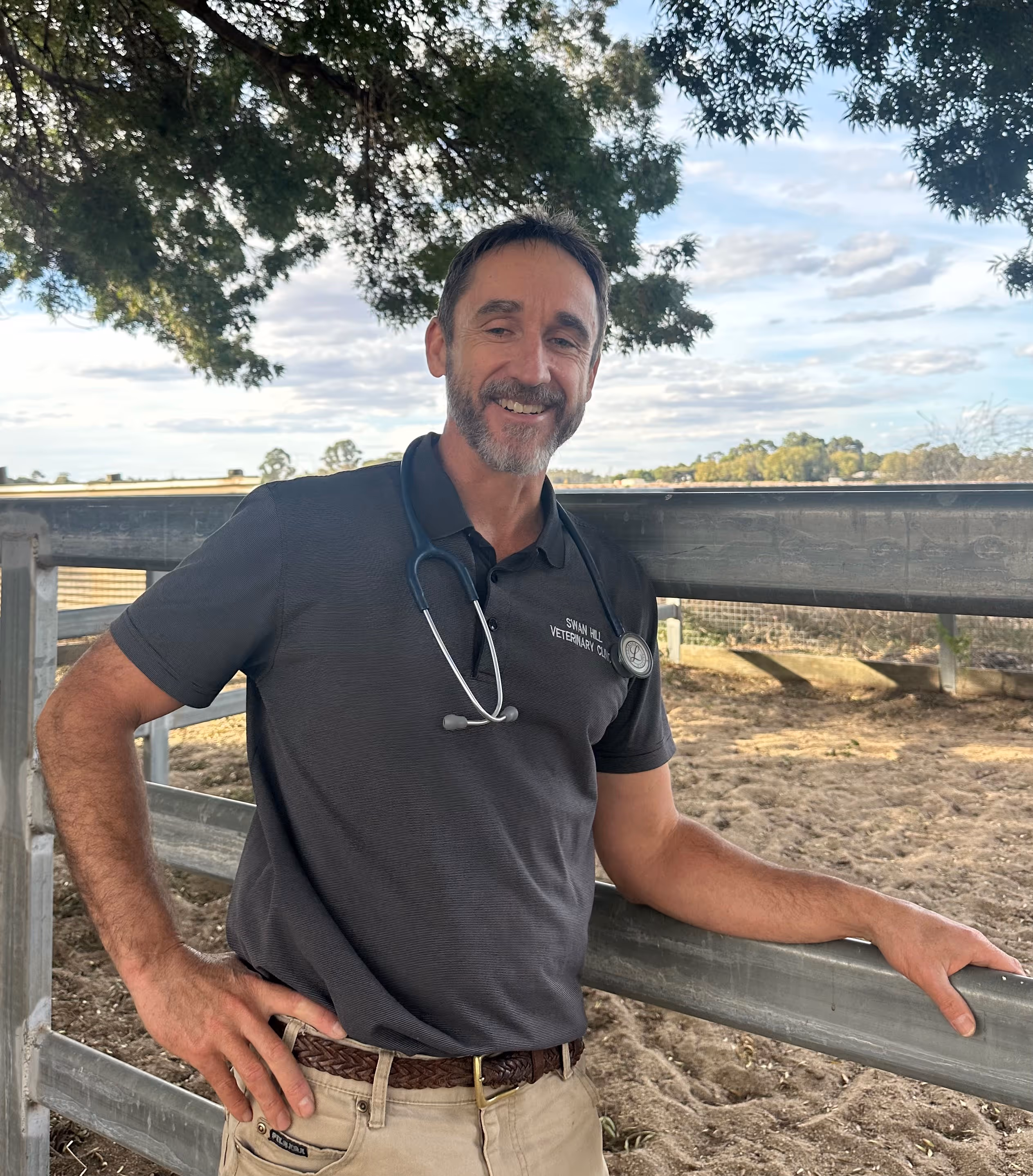 Smiling man with a stethoscope around his neck leaning on a metal fence in an outdoor rural setting.