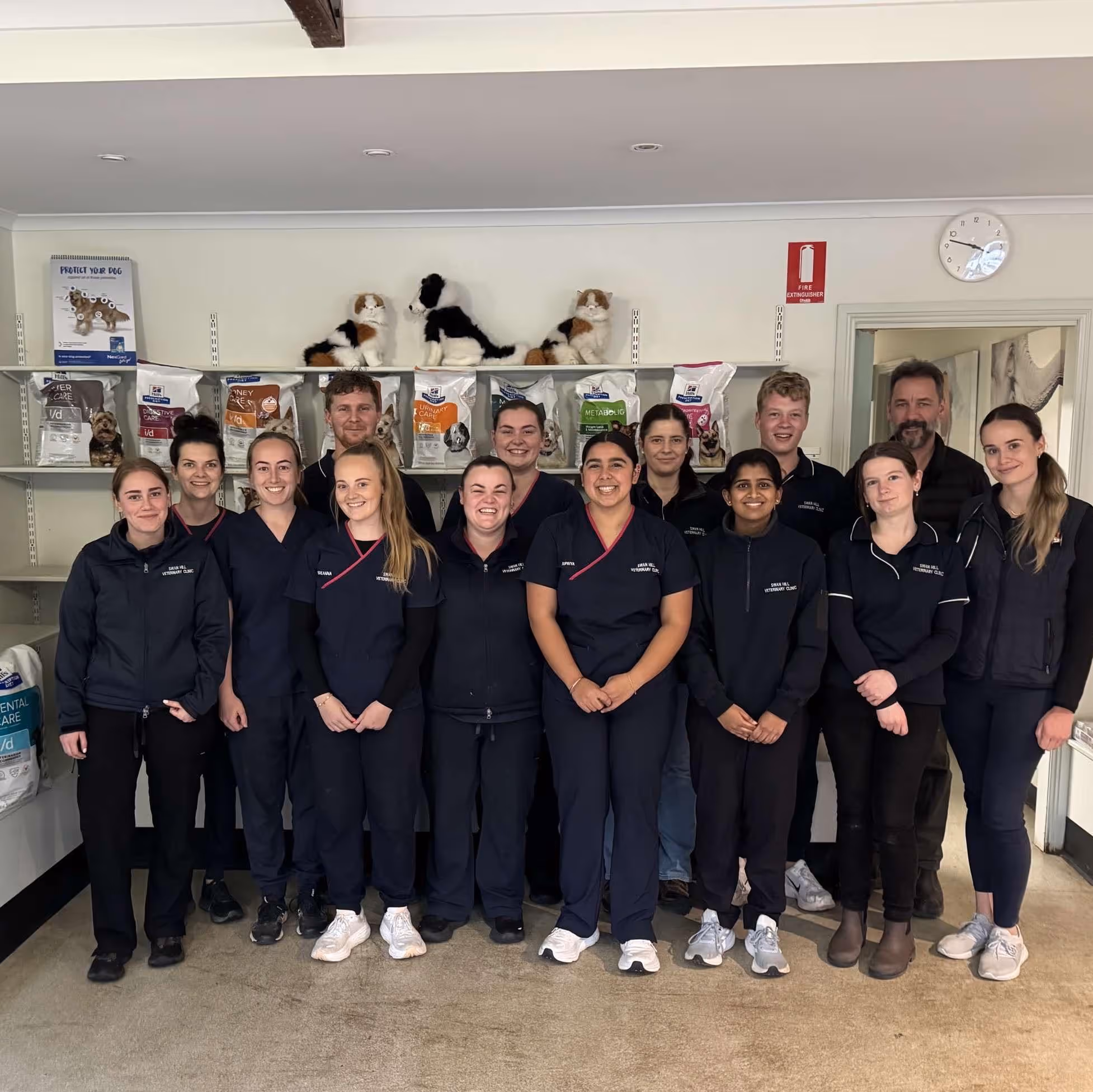 Group photo of 14 veterinary clinic staff members wearing navy uniforms standing indoors with shelves of pet food and stuffed dog toys behind them.