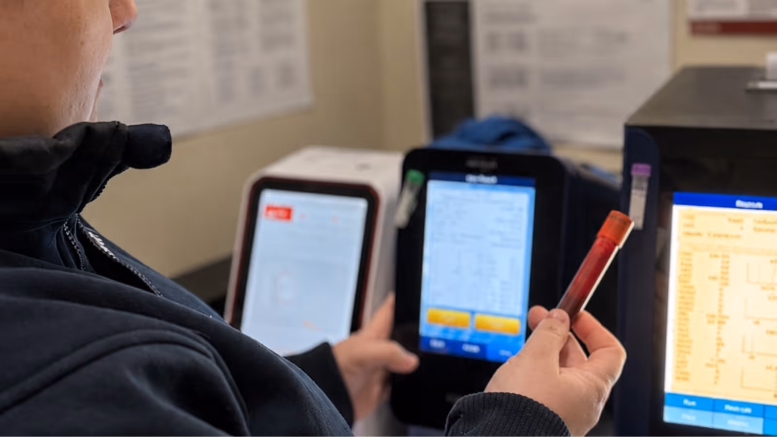 Person holding a blood sample tube near laboratory testing machines with digital screens.