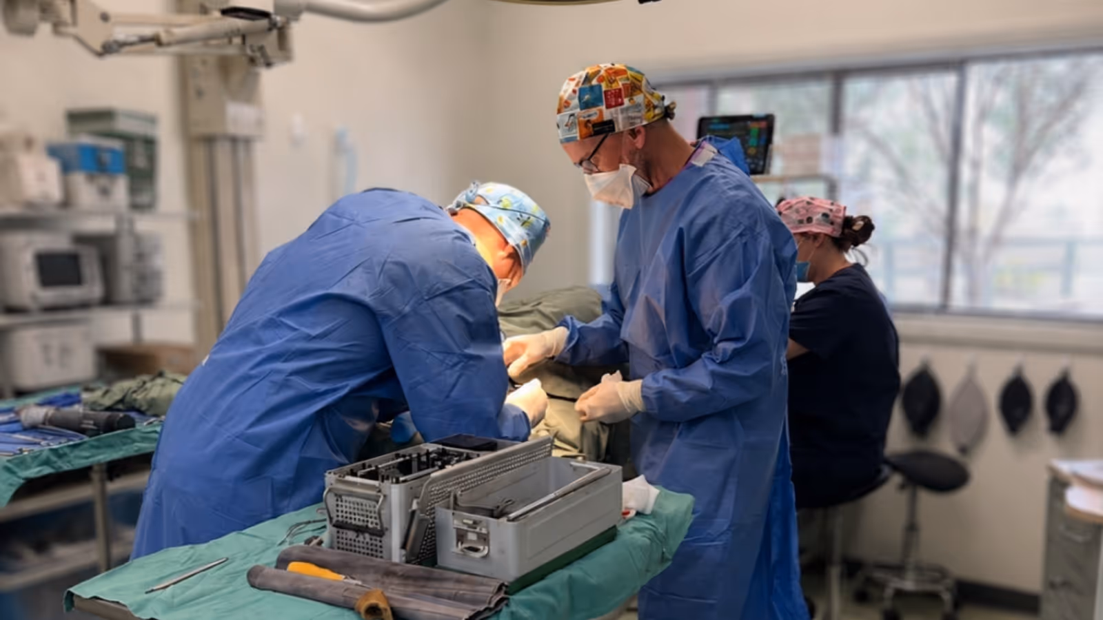 Two surgeons in blue scrubs performing surgery with assistant seated nearby in operating room.