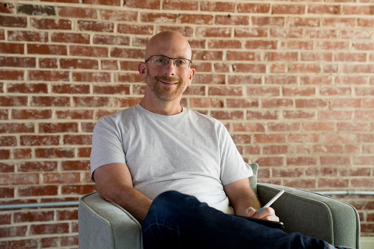 A photo of Matthew Penner sitting in a chair in front of a brick wall holding an iPad and Apple Pencil 