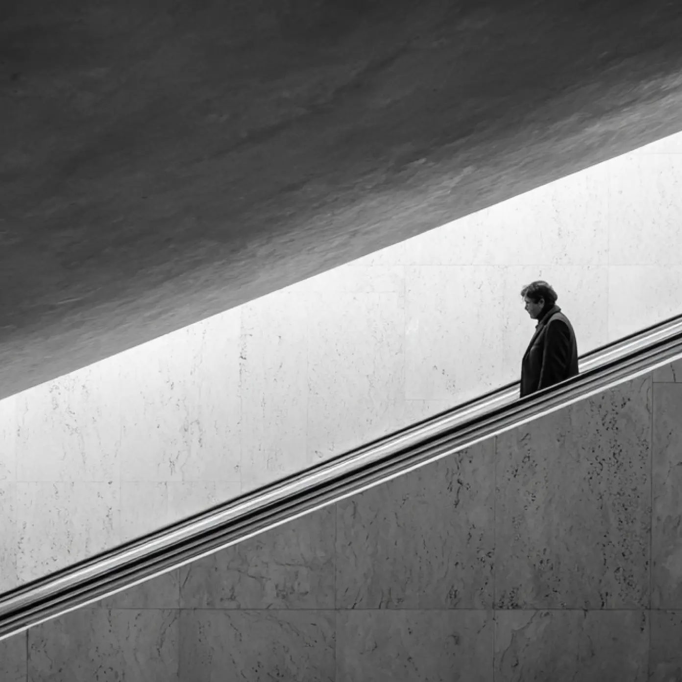 Man with coat riding down an escalator inside a modern building with marble walls.