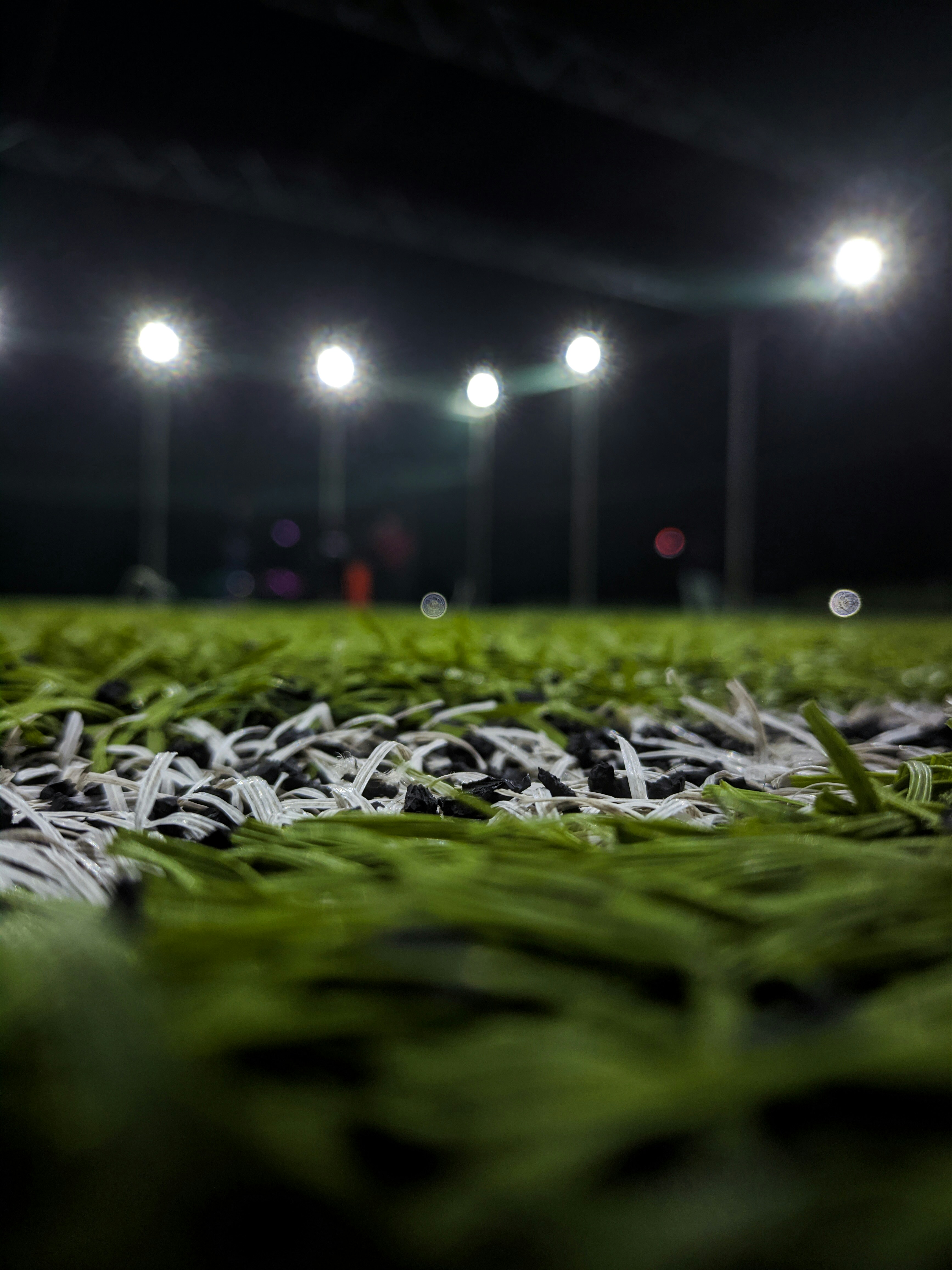 alt="Kunstrasen-Detailaufnahme in Bodenperspektive mit Flutlichtmasten auf beleuchtetem Sportplatz bei Nacht"