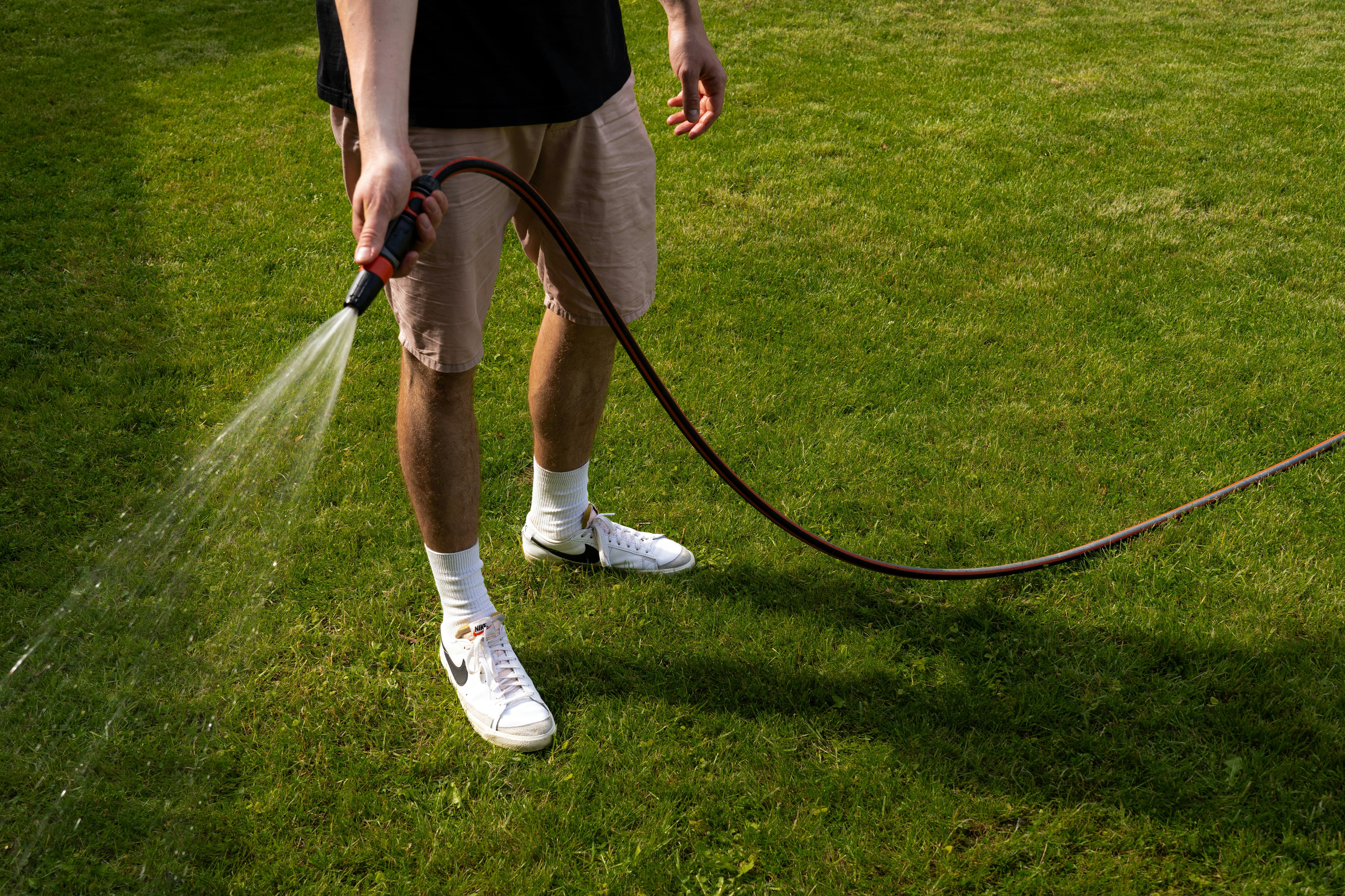 Person wässert einen grünen Rasen mit einem Gartenschlauch und Sprühpistole im Sonnenlicht