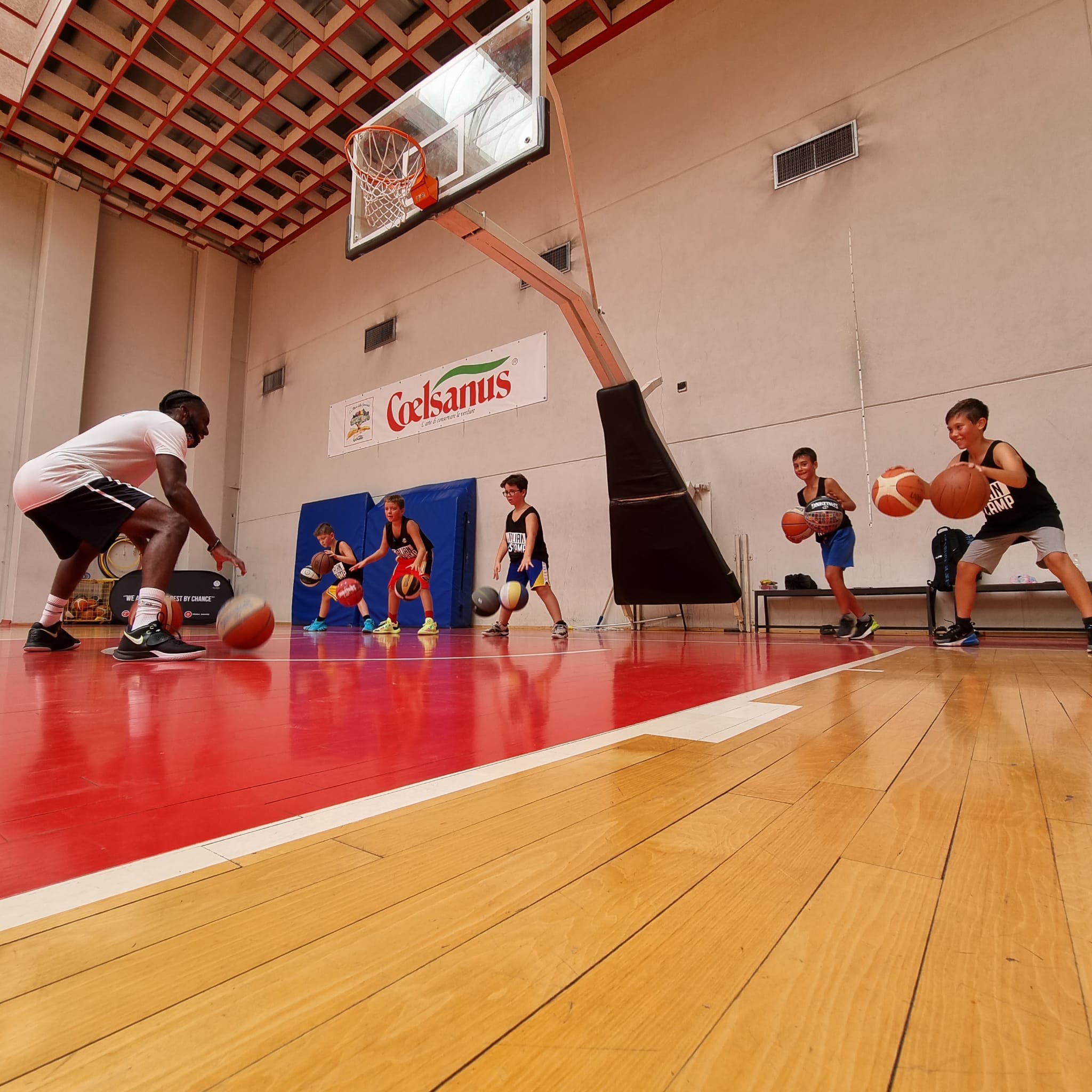 Basketball coach training five kids dribbling balls on an indoor court under a hoop.