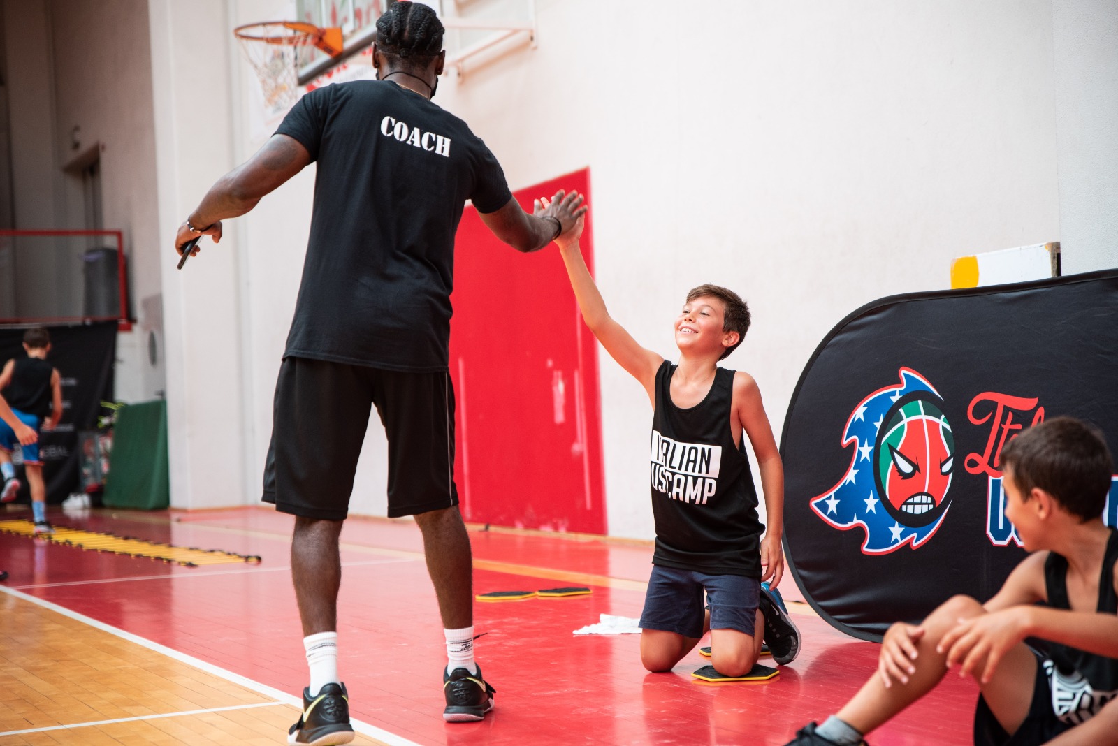 Boy in basketball camp gear high-fives coach during indoor training on red court.