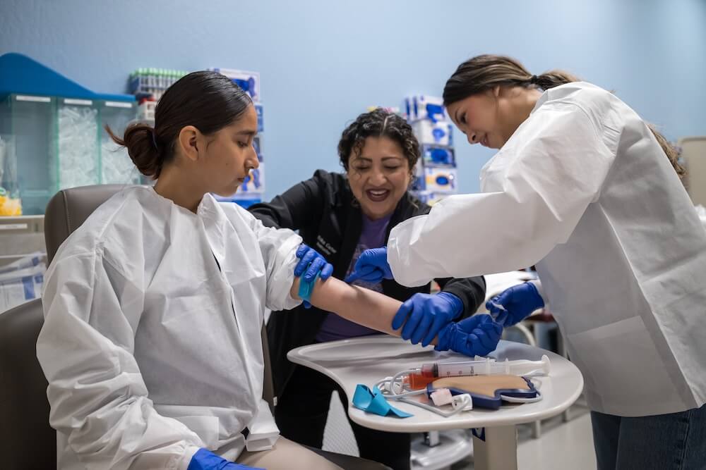 Medical professionals in protective clothing preparing a young woman's arm for a blood draw in a clinical setting.