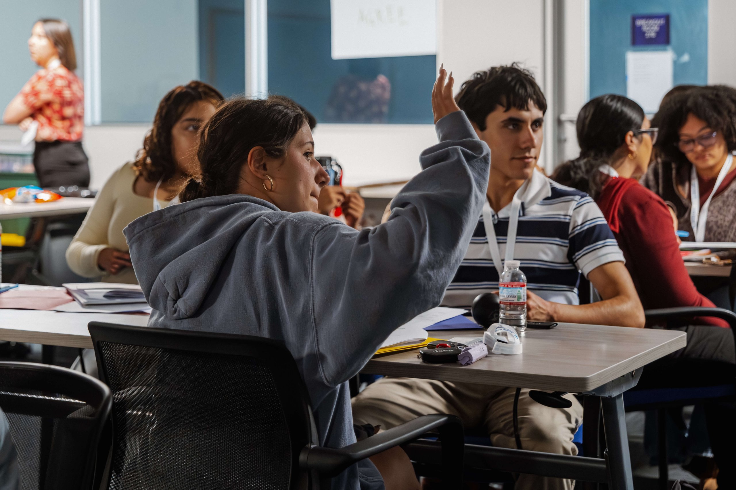 Students sitting in a classroom, one female student raising her hand while others engage in discussion or work.