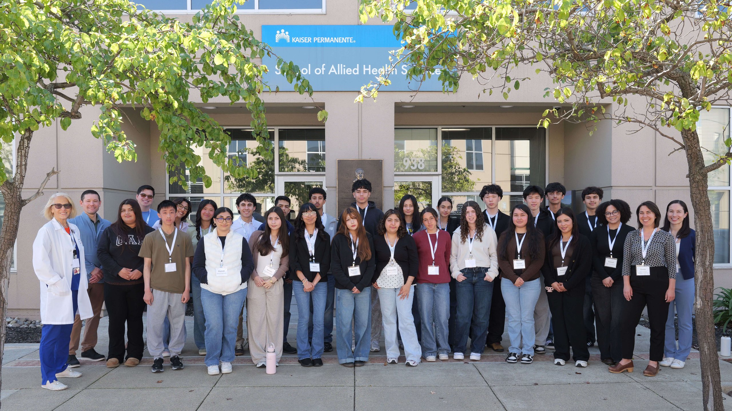 Group photo of diverse students and staff standing outside Kaiser Permanente School of Allied Health Sciences building under trees.