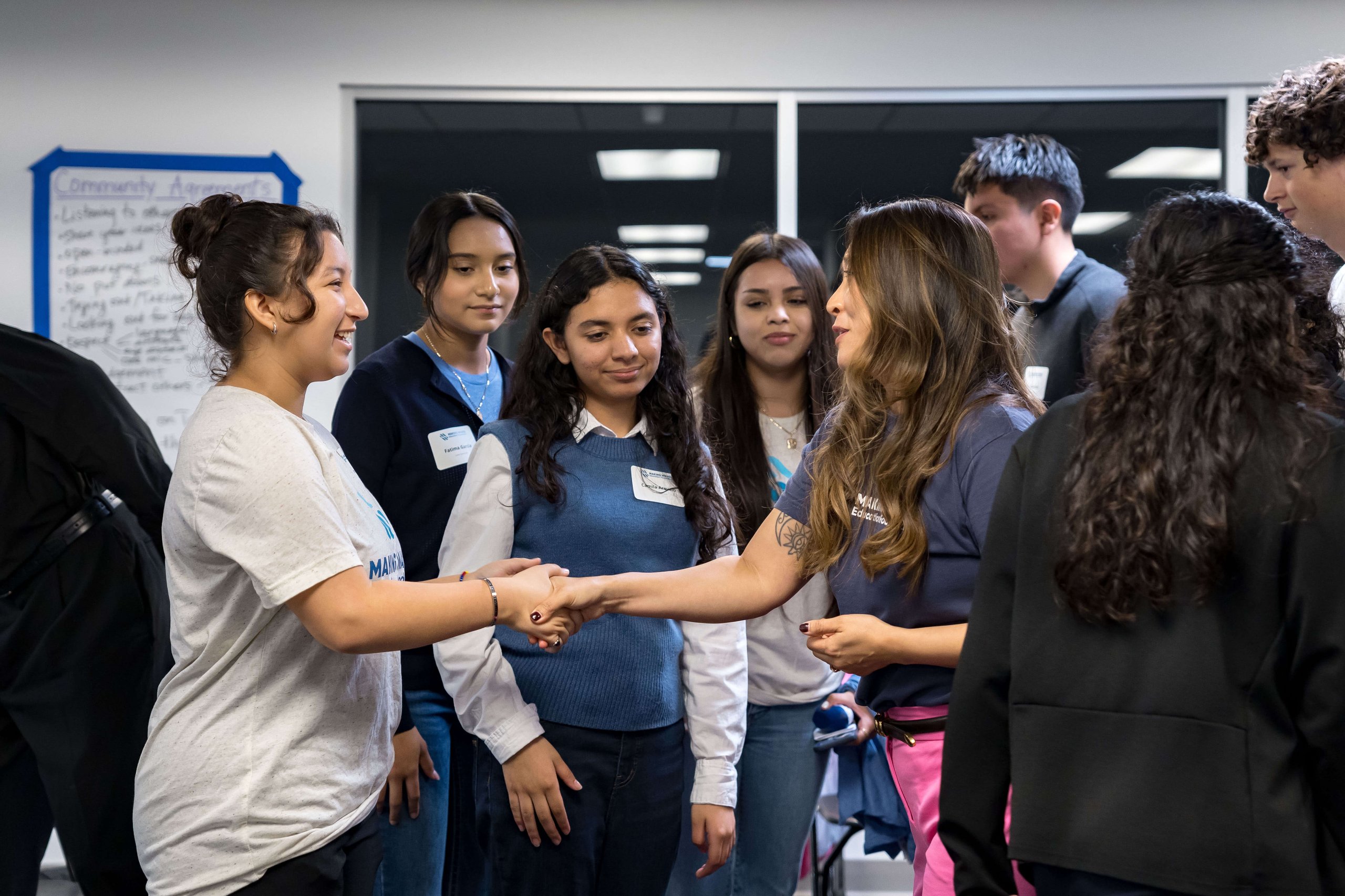 A group of young people indoors, with two women in the foreground shaking hands and others watching.