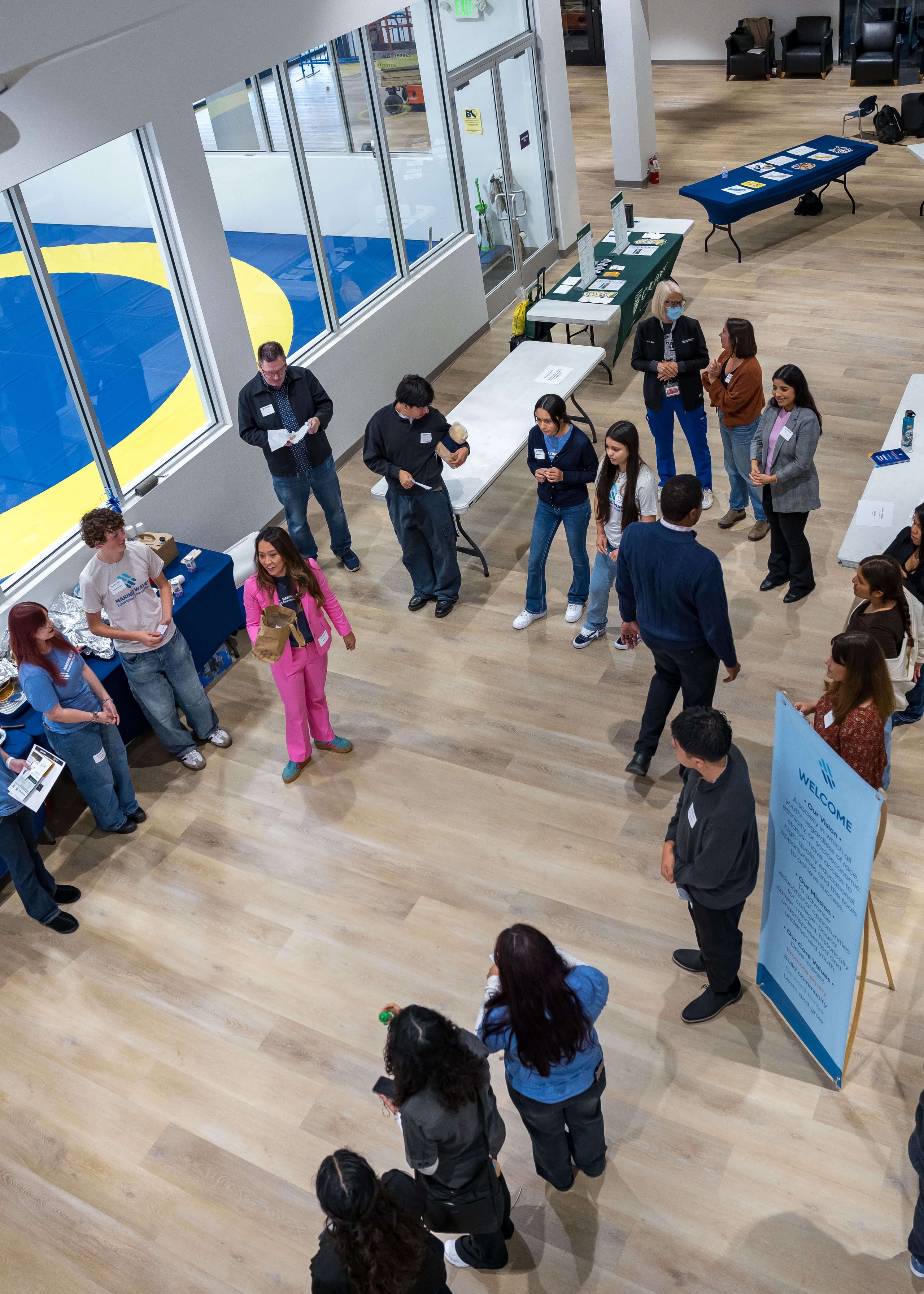 Group of people gathered in a large room with tables displaying informational materials and a visible wrestling mat through windows.