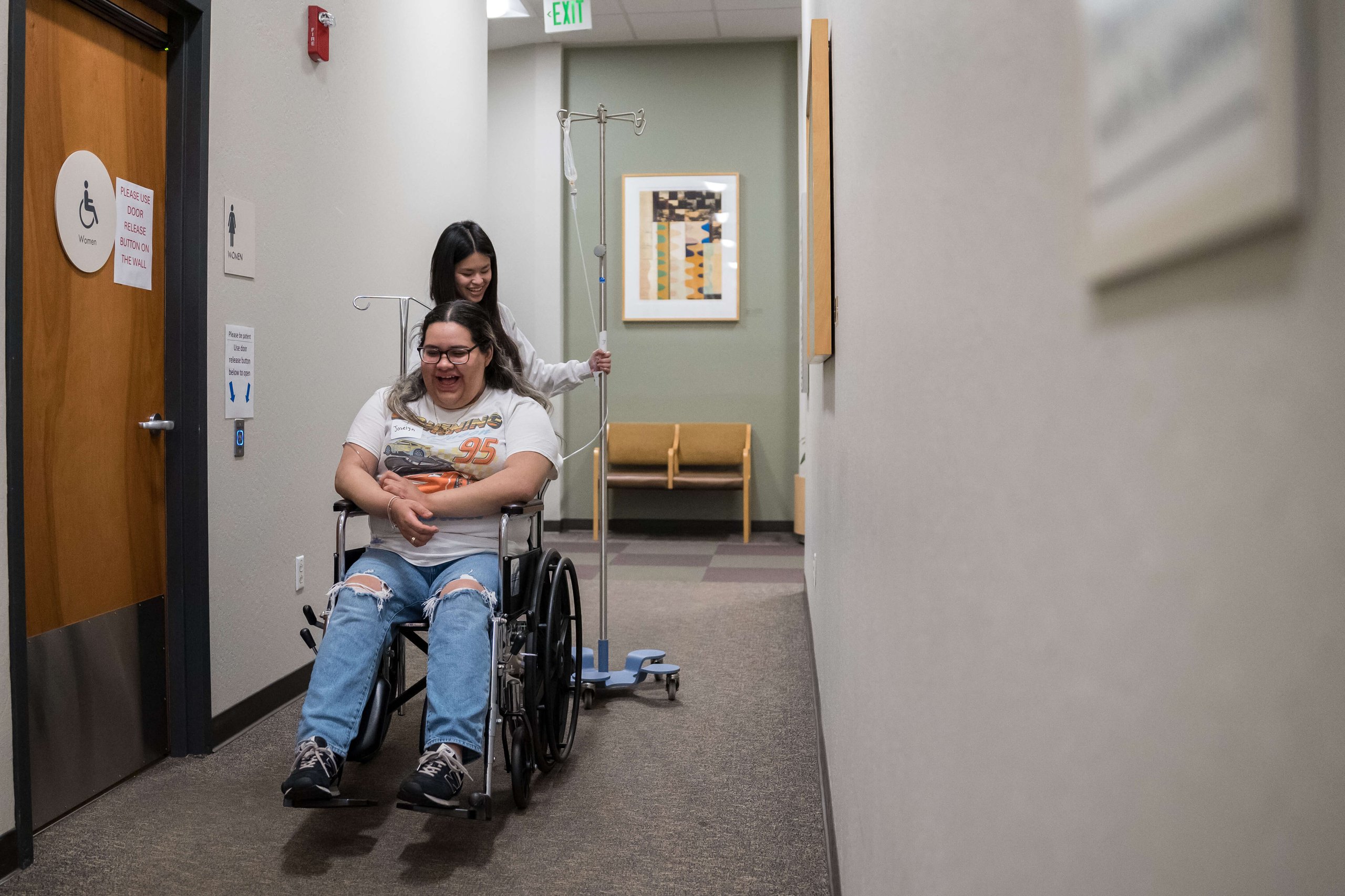 Healthcare worker pushing smiling patient in a wheelchair down a hospital corridor while holding an IV pole.