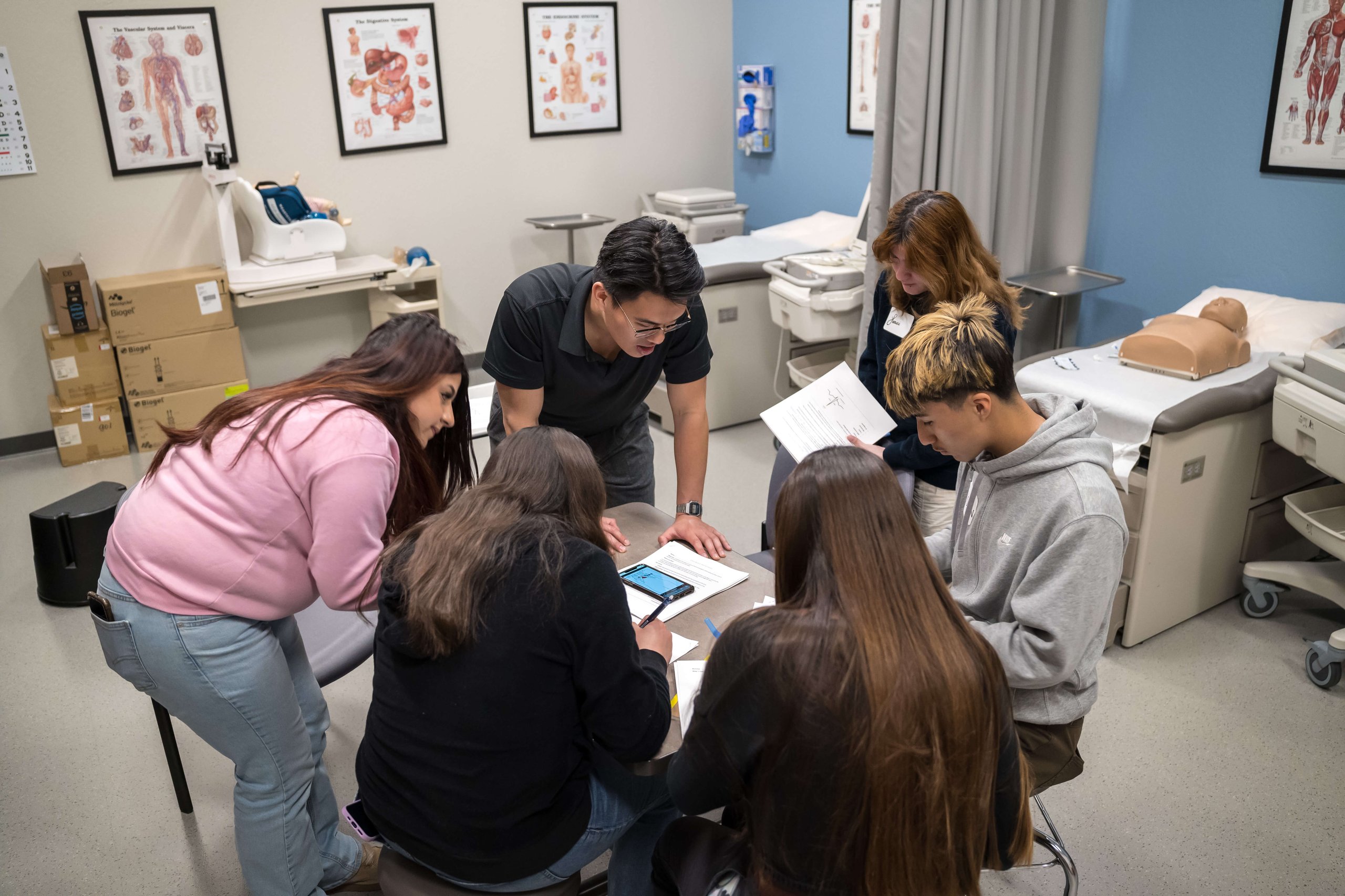 Students gathered around a table reviewing healthcare training materials in a clinical skills classroom.