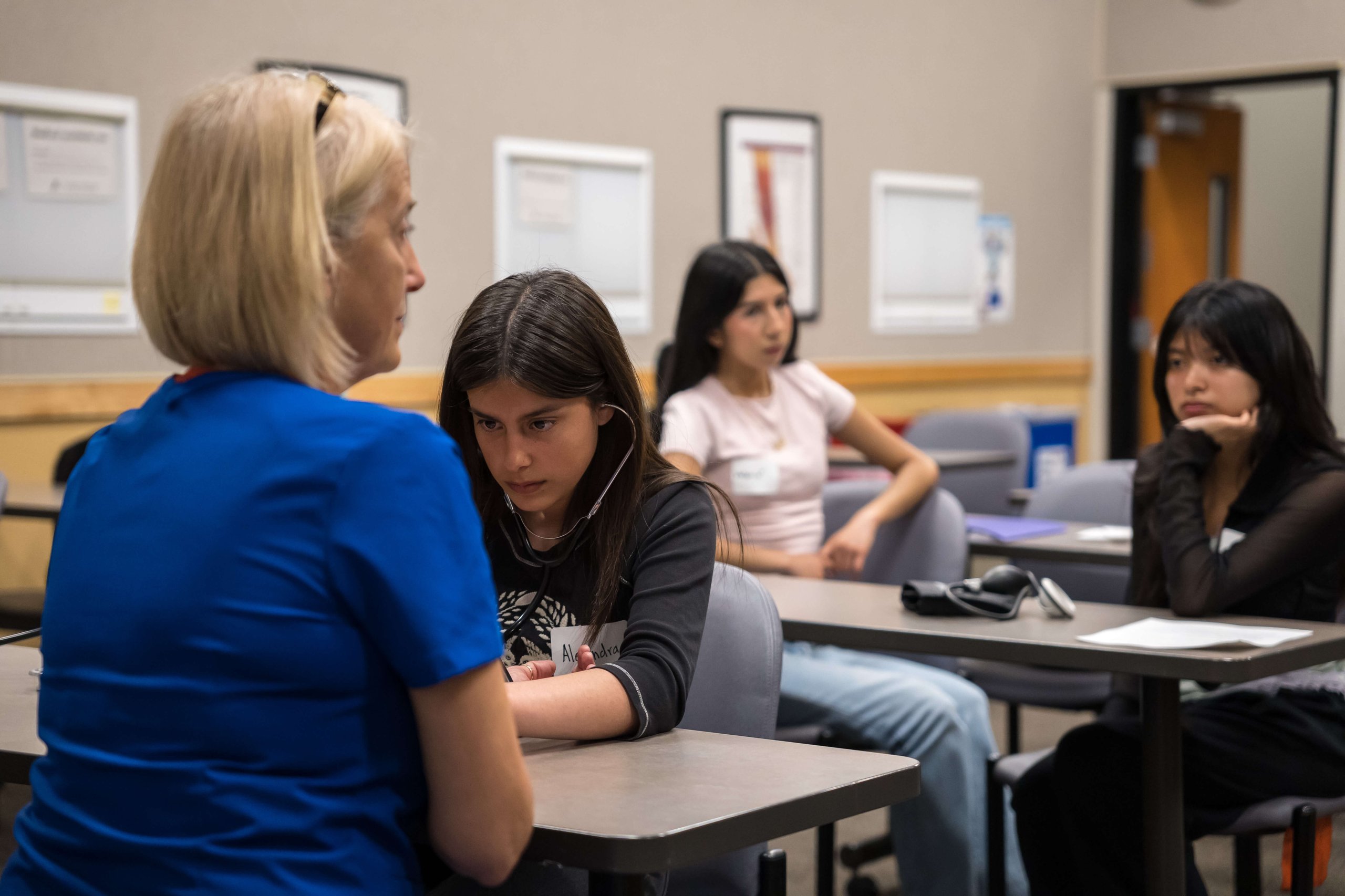 A woman in a blue shirt instructs a young girl using a stethoscope to listen to her arm while two other girls watch in a classroom setting.