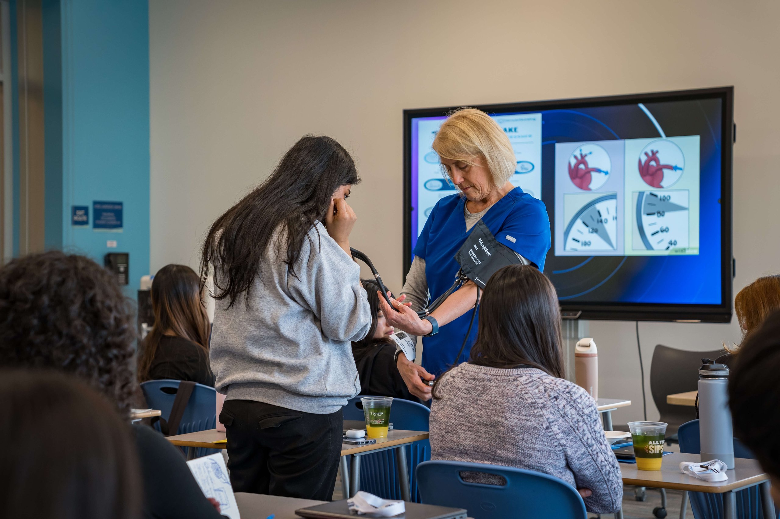 Healthcare instructor in blue scrubs measures student’s blood pressure in classroom with heart diagrams on screen.