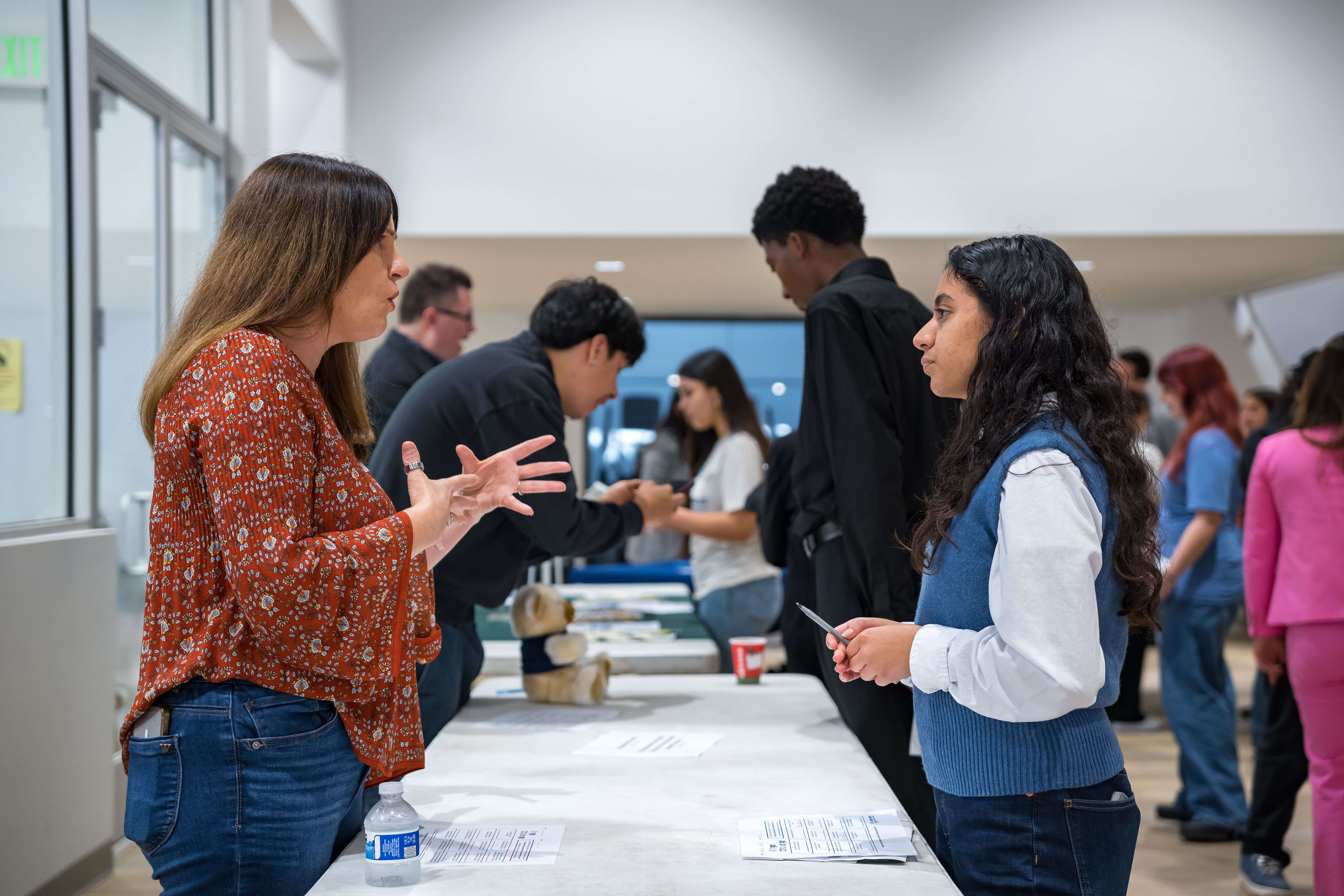 Young people talking to a woman in a gray blazer at a table with informational papers in a bright indoor space.