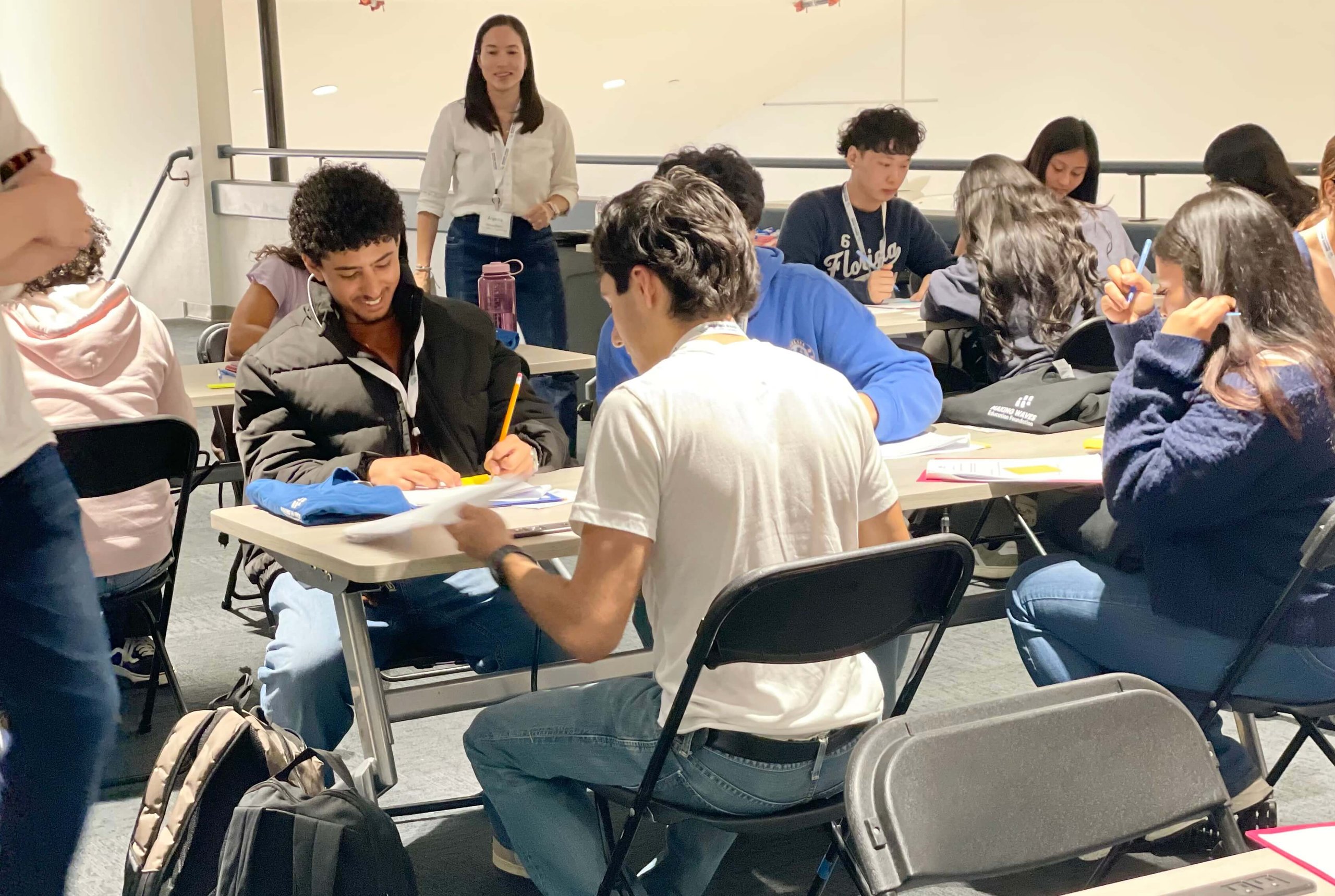 Students seated at tables working on papers, with a standing teacher observing in a classroom setting.