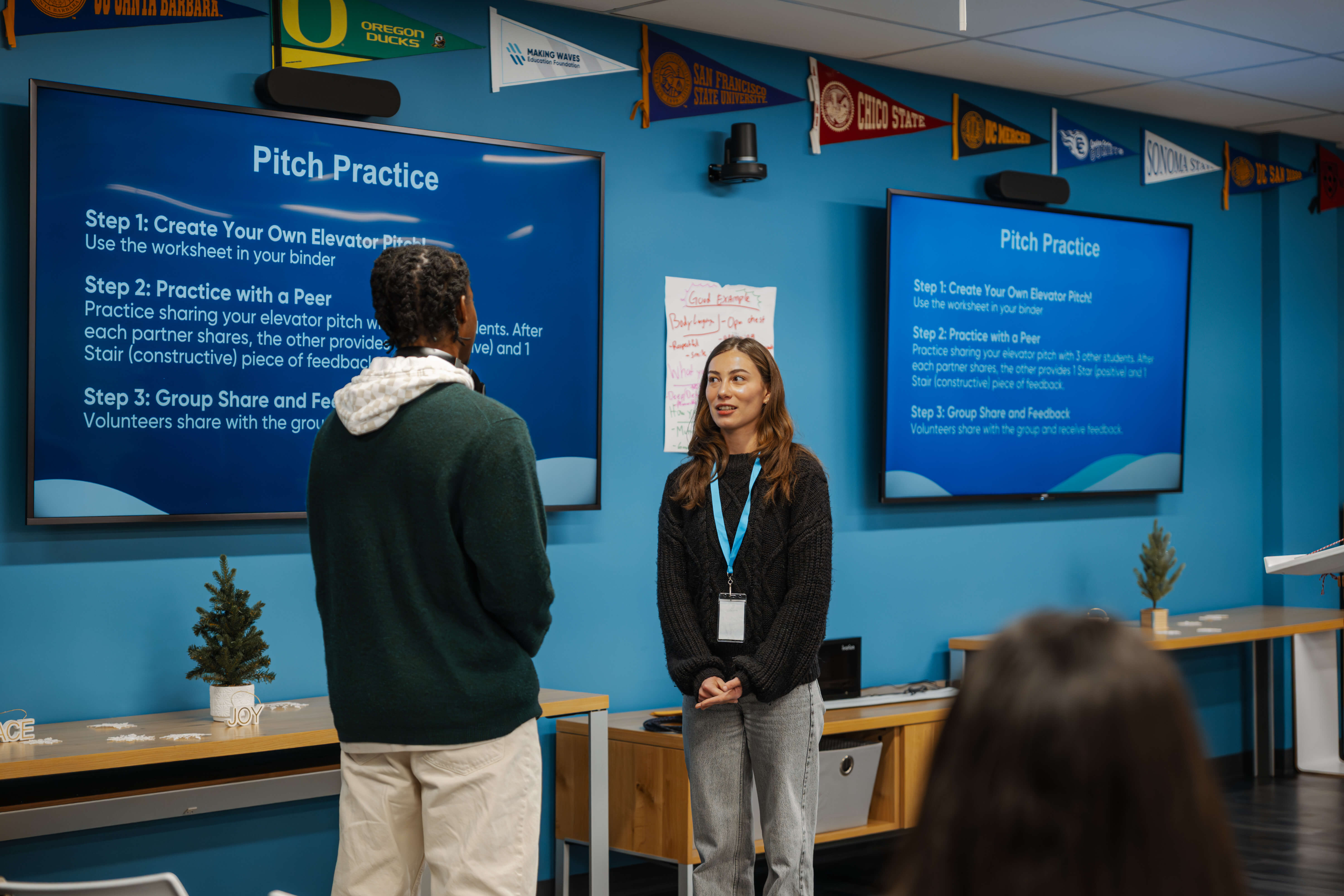 Two people practicing elevator pitches in a blue classroom with pitch practice instructions on screens.