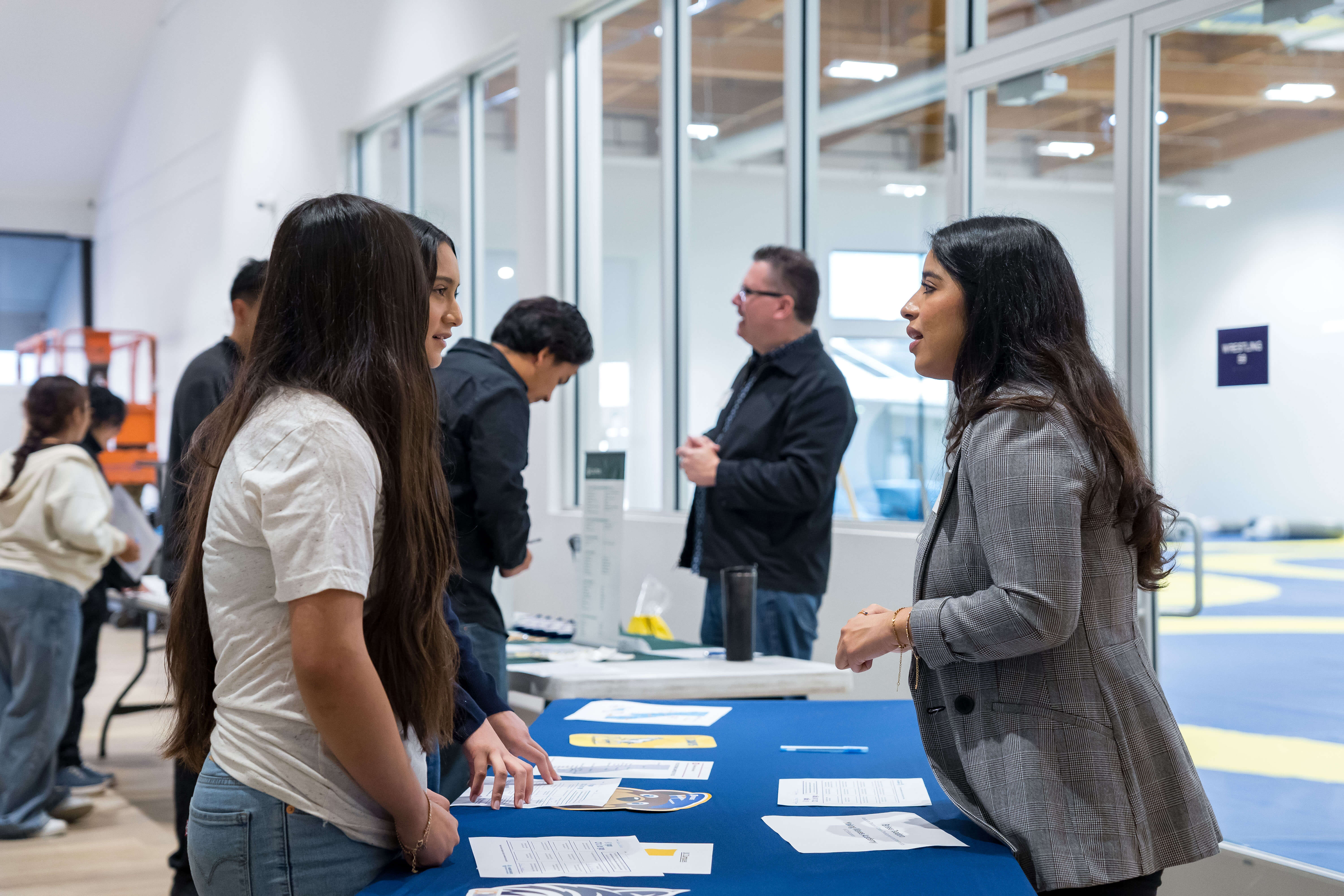 Two young women at a job fair table speaking with a recruiter in a gray blazer indoors.