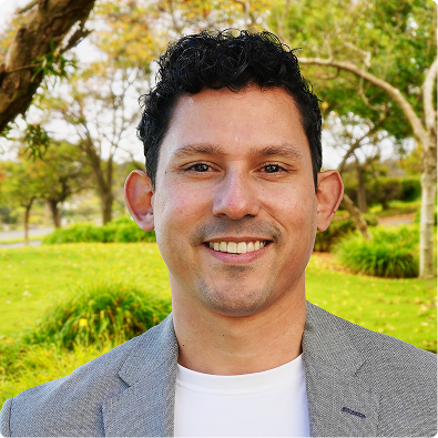 Smiling man with curly dark hair wearing a gray blazer and white shirt in a park.