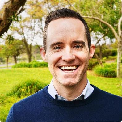 Smiling man with short dark hair wearing a navy sweater and white collared shirt outdoors in a park.