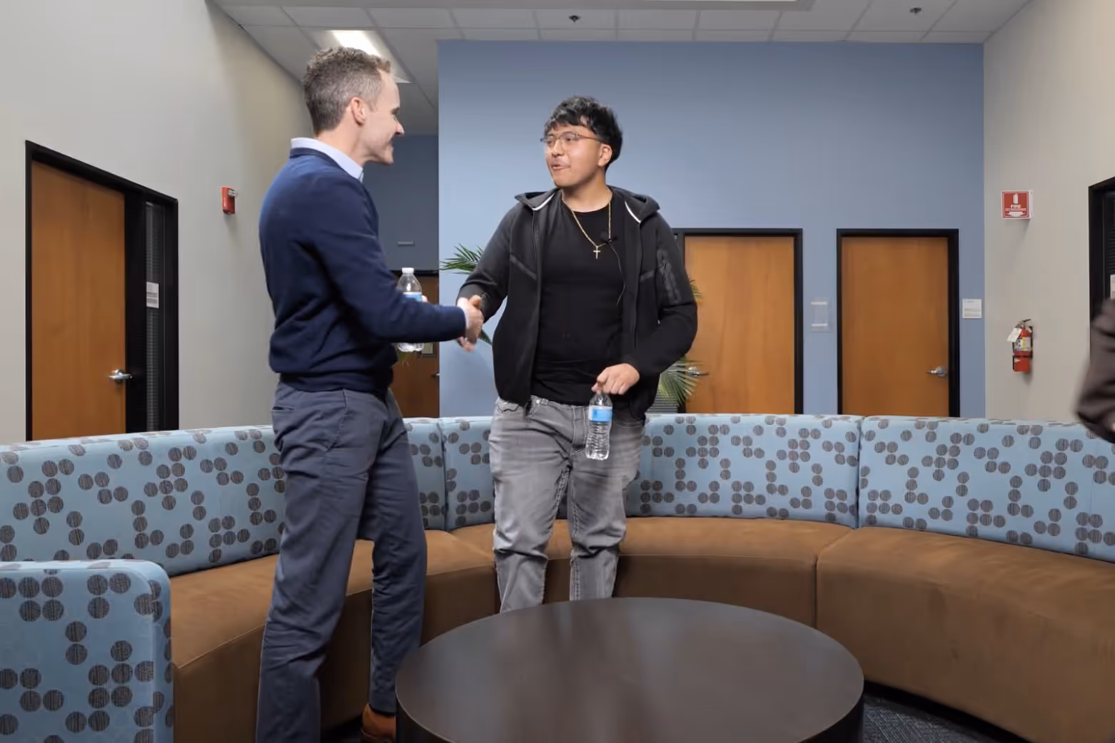 A professional man in a navy blue sweater and grey slacks smiles while shaking hands with a young man in glasses and a black hoodie inside a modern office lobby with a large curved couch.