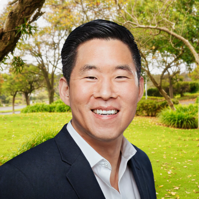 Smiling man in a white shirt and dark blazer standing in a green park with trees.