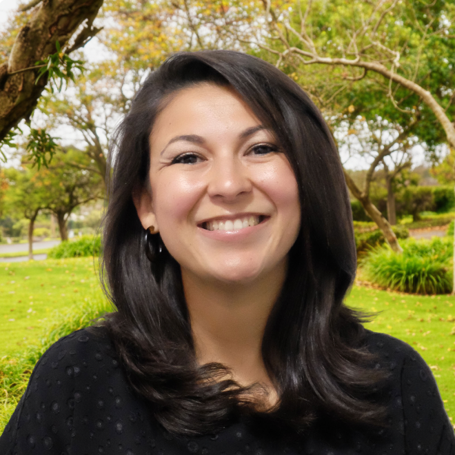 Smiling woman with dark hair standing outdoors with green trees and grass in background.