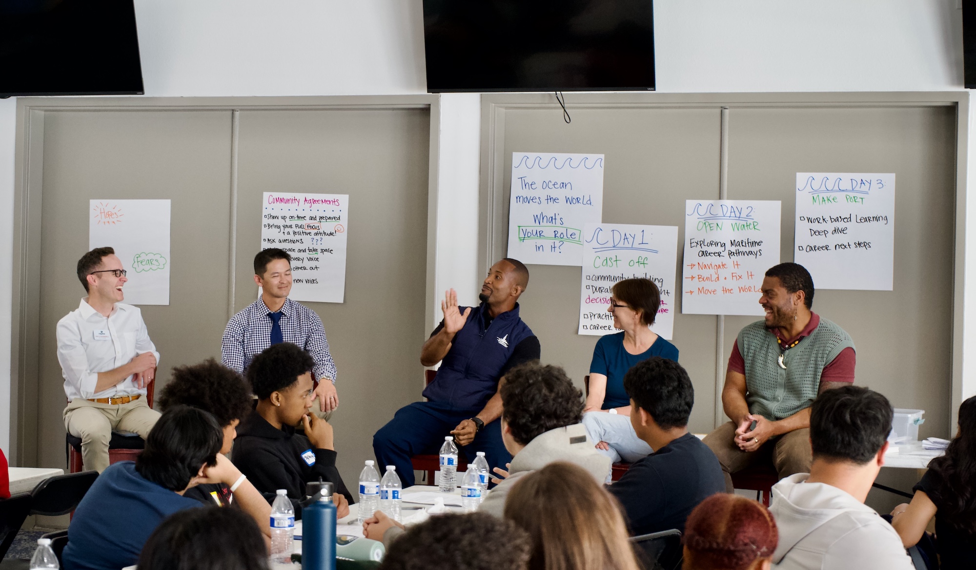 Panel of five people talking in front of an audience with posters about maritime career pathways behind them.