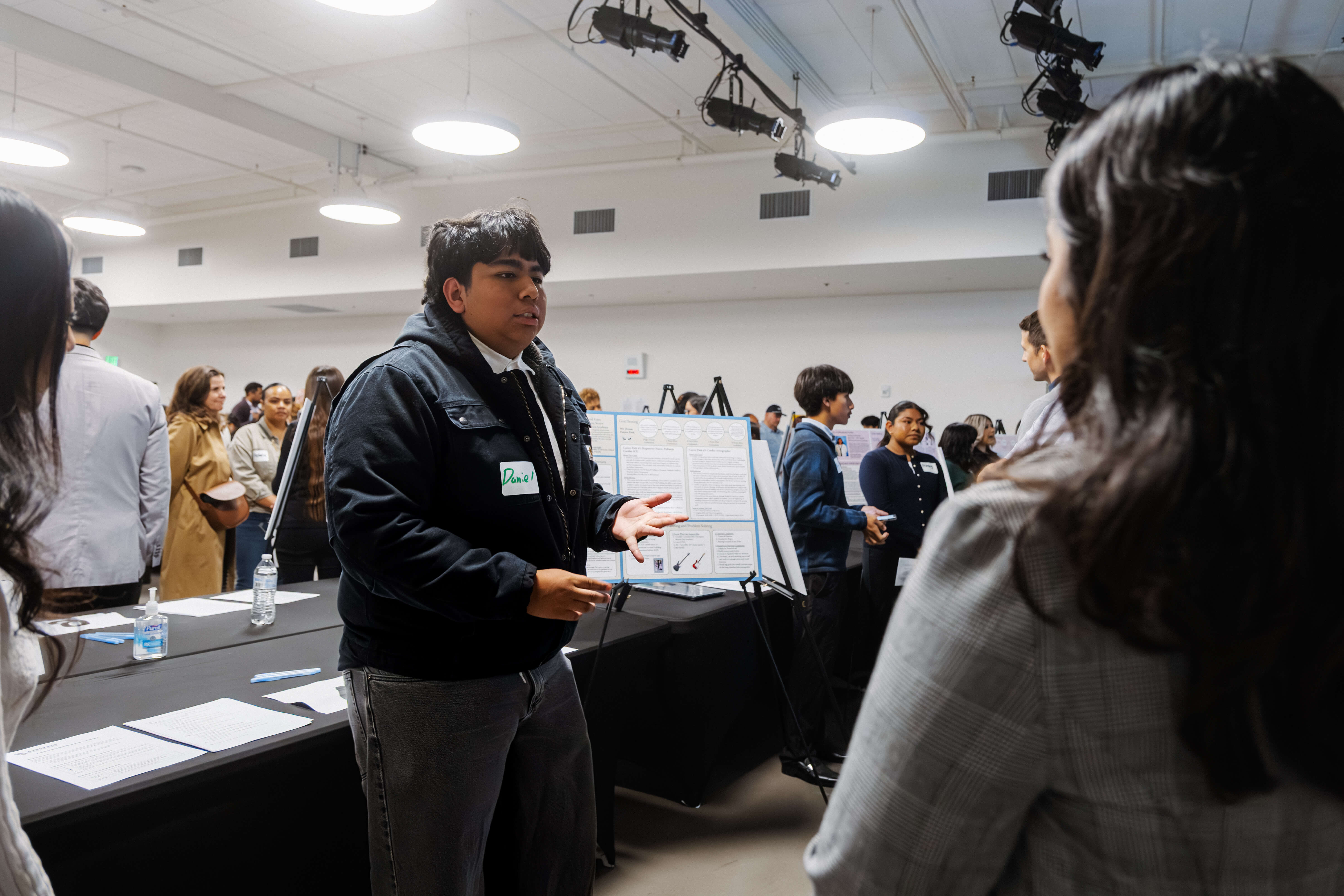 A young man wearing a black jacket with a name tag saying 'Daniel' is presenting information on a poster board in a room with several other people and presentation boards visible in the background.