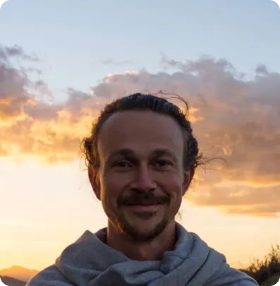Smiling man with curly hair and a gray hoodie standing outdoors with a sunset sky background.
