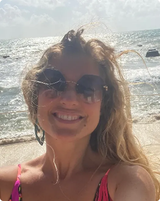 Smiling woman with sunglasses and wavy hair taking a selfie on a sunny beach with sparkling water.