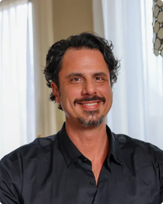 Smiling man with dark hair and goatee wearing a black shirt in a softly lit room.