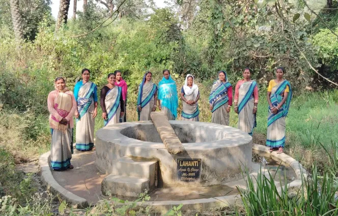 Ten women in saris standing around a traditional water well in a green forested area.