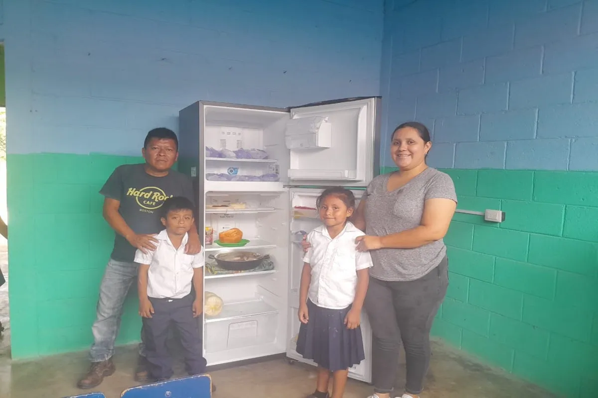 Family of four stands inside a room beside an open refrigerator with food inside.