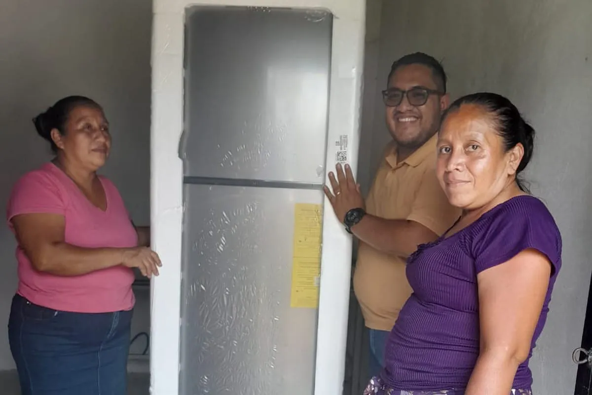 Three people smiling next to a silver refrigerator wrapped in protective foam inside a bare room.