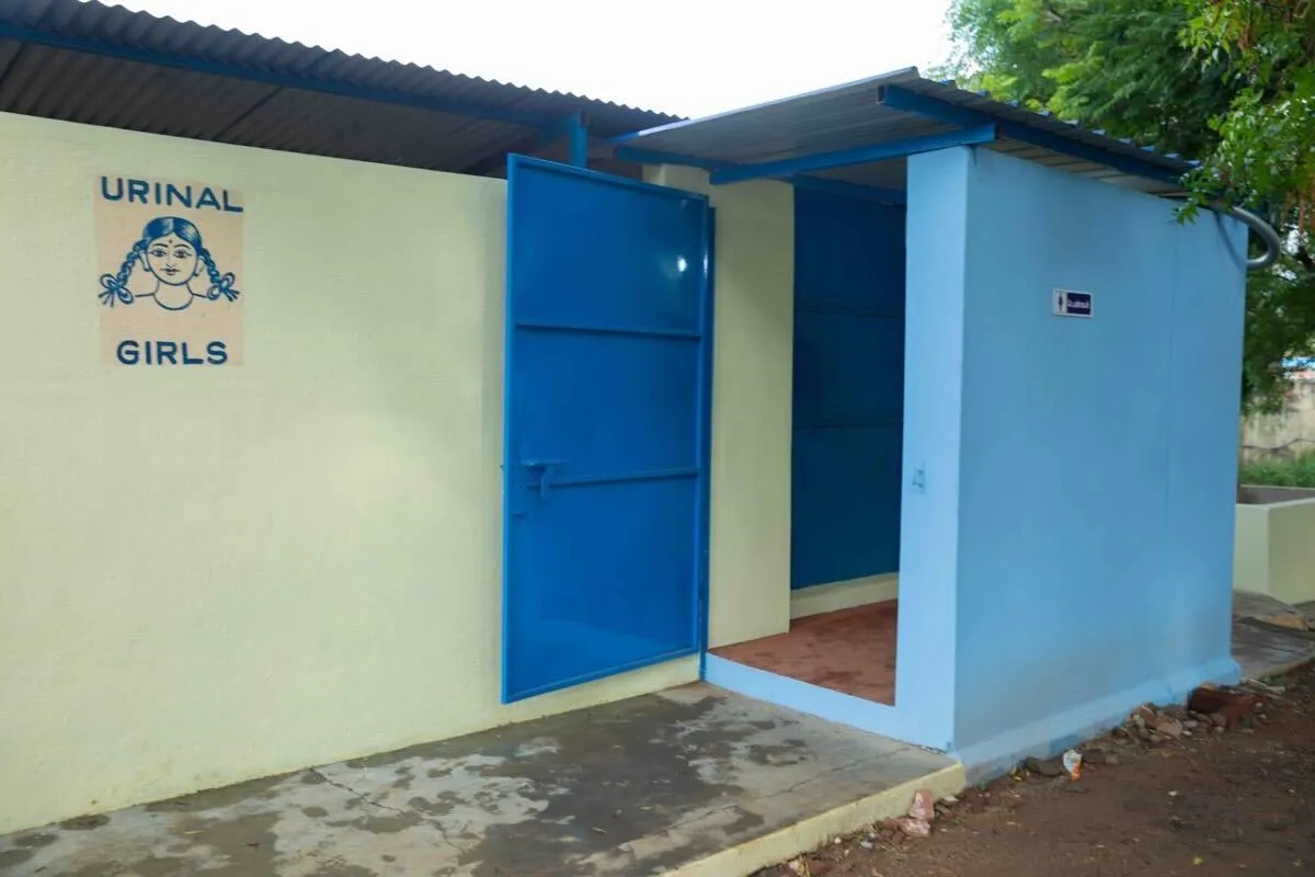 Outdoor latrine with a drawing of a girl and the words URINAL GIRLS on the wall.