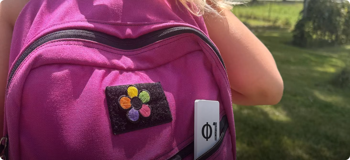 Close-up of a child's purple backpack with a flower patch and a white Phi device in the zippered pocket.