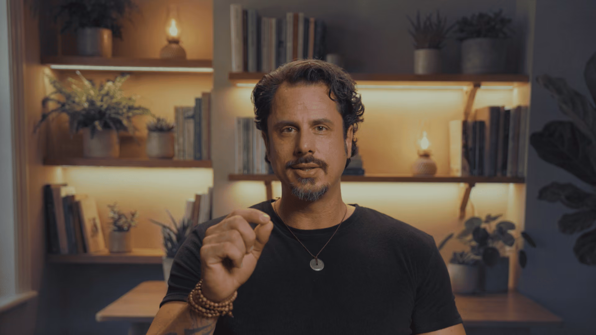 Man with short curly hair and beard gesturing with fingers in a warmly lit room with shelves and plants.